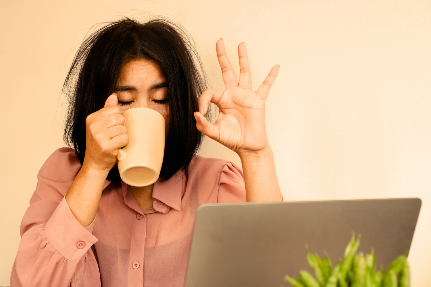 Transparente Tired woman sitting in front of a computer drinking out of a mug and holding up hand in an “I’m ok” symbol.